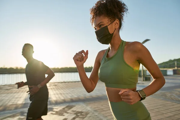 Young black sports couple running on river promenade in city. Slim woman and athletic man wear sportswear snd medical masks. Healthy lifestyle. Health protection during Coronavirus pandemic. Sunny day