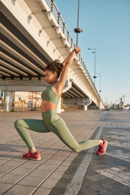 Side view of young serious black sportswoman stretching on blurred city area. Pretty slim curly girl wearing sportswear and looking away. Concept of modern healthy female lifestyle. Sunny daytime