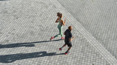 Top view of young black sports couple running on empty city area. Attractive slim curly girl and athletic man wearing sportswear. Concept of modern healthy lifestyle. Warm sunny day