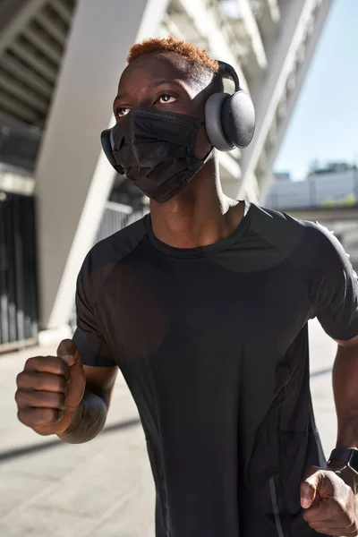 Young black sportsman running near blurred stadium. Guy wearing sportswear, smartwatch, headphones and medical mask. Modern healthy lifestyle. Health protection during COVID-19 epidemic. Sunny day