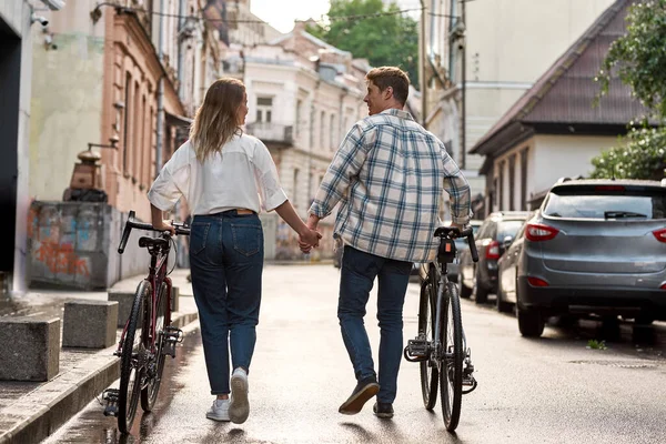 Back view of young couple walking with bicycles on city street. Modern urban healthy lifestyle. Entertainment, leisure and hobby. Relationship. Man and girl holding hands and looking at each other