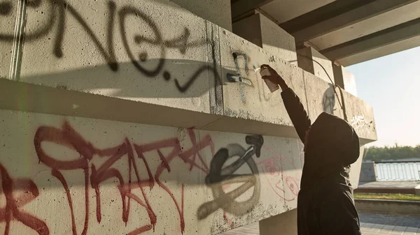 Black man drawing graffiti letters and symbols with aerosol can on concrete building in city. Street art. Hip hop culture. Male wearing hoodie. Modern urban lifestyle. Sunny day