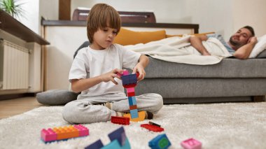 Babası evdeki kanepede uyurken inşaat bloklarıyla oynayan küçük bir çocuk. Genç, modern, beyaz aile yaşam tarzı ve ilişkisi. Ev eğlencesi, hobi ve eğlence