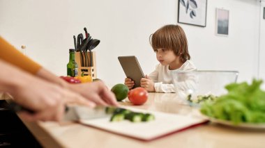 Küçük beyaz çocuk dijital tablet kullanırken annesi evde salata pişirmek için salatalık doğruyor. Annelik ve ebeveynlik. Aile ilişkisi. Yurt içi yaşam tarzı