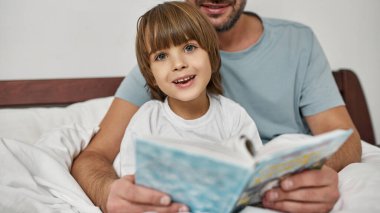 Babası evde ona sarılıp kitap okurken küçük çocuk kameraya bakıyor. Genç, beyaz bir aile ilişkisi. Babalık ve ebeveynlik. Ev eğlencesi, hobi ve eğlence