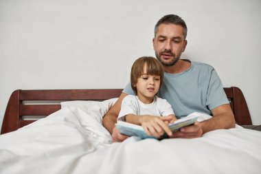 Babası evde küçük oğluna sarılıp kitap okuyordu. Genç, beyaz bir aile ilişkisi ve birlikte vakit geçirmek. Babalık ve ebeveynlik. Ev eğlencesi, hobi ve eğlence