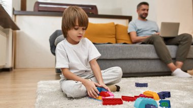Babam evdeki koltukta dizüstü bilgisayar kullanırken küçük çocuk halıda inşaat taşlarıyla oynuyor. Modern Kafkas aile yaşam tarzı ve ilişkisi. Ev eğlencesi, hobi ve eğlence