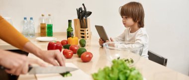 Beyaz çocuk dijital tablet kullanırken, annesi evde salata pişirmek için salatalık doğruyor. Annelik ve ebeveynlik. Aile ilişkisi. Yurt içi yaşam tarzı