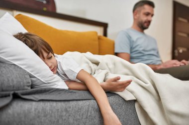 Evdeki kanepede bulanık babasının yanında battaniyenin altında uyuyan küçük çocuğun ön planına odaklan. Genç, beyaz bir aile ilişkisi. Babalık ve ebeveynlik. Modern yerli yaşam tarzı