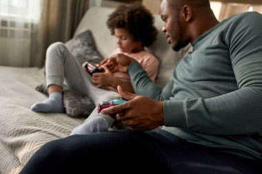 Siyah baba evdeki kanepede video oyunu oynarken bulanık oğlunun joystick 'ini itiyor. Küçük çocuğa yardım eden adam. Babalık ve ebeveynlik. Ev içi hobi, eğlence ve eğlence.