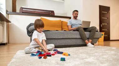 Küçük çocuk halıda inşaat bloklarıyla oynarken babası evdeki kanepede dizüstü bilgisayar izliyor. Genç, modern, beyaz aile yaşam tarzı ve ilişkisi. Ev eğlencesi, hobi ve eğlence