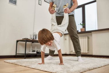 Evdeki halının üzerinde duran küçük oğlunun bacaklarını tutan kısmi baba. Genç bir aile ilişkisi ve birlikte zaman geçirmek. Babalık ve ebeveynlik. Sağlıklı bir ev hayatı. Boş zaman