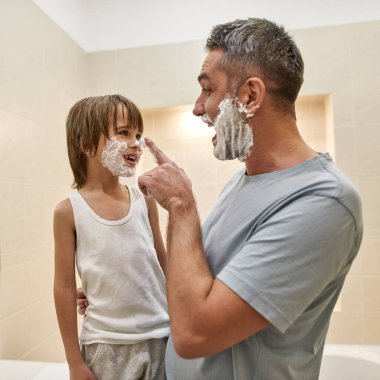 Neşeli baba, tıraş olurken gülümseyen küçük oğlunun burnuna tıraş köpüğü sürüyor. Beyaz çocuk ve erkek şakalaşıyor ve birlikte vakit geçiriyorlar. Babalık. Yüz bakımı ve hijyen