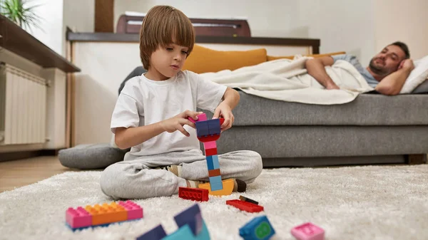 Babası evdeki kanepede uyurken inşaat bloklarıyla oynayan küçük bir çocuk. Genç, modern, beyaz aile yaşam tarzı ve ilişkisi. Ev eğlencesi, hobi ve eğlence