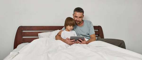 Εστιασμένος Πατέρας Και Μικρός Γιος Βλέποντας Ψηφιακή Πλακέτα Στο Κρεβάτι — Φωτογραφία Αρχείου