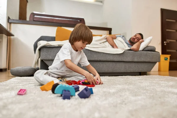 父親が家でソファで寝ている間 小さな男の子はカーペットの上の建設ブロックで遊んでいます 若い現代ヨーロッパの家族のライフスタイルと関係 国内レジャー — ストック写真