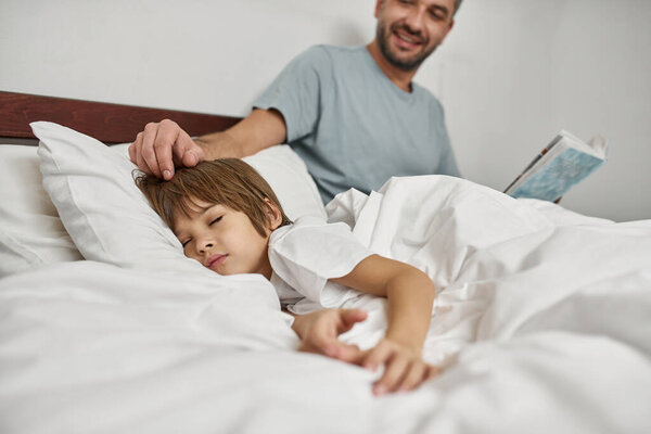 Smiling father with book caressing his sleeping little son in bed at home. Family relationship and spend time together. Young caucasian boy and man. Fatherhood and parenting. Modern domestic lifestyle