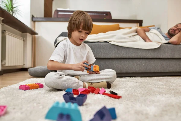 父親が家でソファで寝ている間 小さな男の子はカーペットの上の建設ブロックで遊ぶ 若い現代の白人の家族生活と関係 国内レジャー — ストック写真