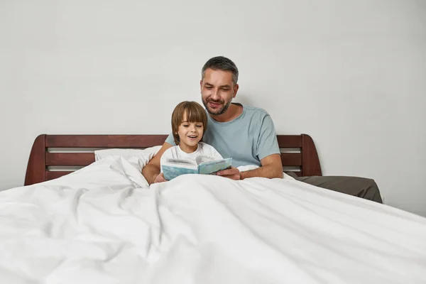 お父さんは家でベッドで小さな息子に本を抱きかかえて読んでいます 若いヨーロッパの家族関係と一緒に時間を過ごす 父親と子育て 国内レジャー — ストック写真
