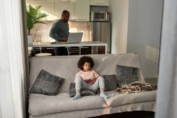 Yetişkin babası evde dizüstü bilgisayar kullanırken ve izlerken konsantre küçük çocuk kanepede joysticklerle video oyunu oynuyor. Siyah adam ve çocuk. Ev içi eğlence ve eğlence.