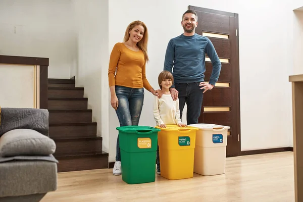 Young happy caucasian family standing near three dustbins for different trash at home. Ecological garbage recycle. Parenting. Father, mother and little son looking at camera. Modern domestic lifestyle