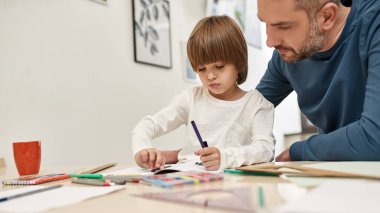 Odaklanmış baba, evdeki masada keçeli kalemle çizim yapan oğluna bakıyor. Beyaz adam ve küçük çocuk ilişkisi. Babalık ve ebeveynlik. Ev eğlencesi, hobi ve eğlence