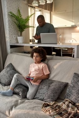 Yetişkin babası evde dizüstü bilgisayarla masada akıllı telefon kullanırken küçük çocuk kanepede joysticklerle video oyunu oynuyor. Siyah adam ve çocuk. Ev içi eğlence ve eğlence.