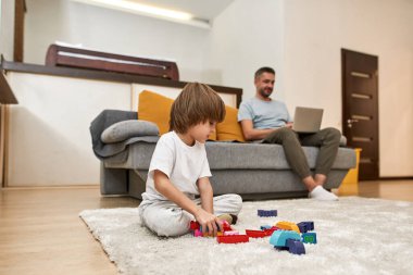 Küçük çocuk halıda inşaat bloklarıyla oynarken bulanık baba evdeki kanepede dizüstü bilgisayar kullanıyor. Genç, modern, beyaz aile yaşam tarzı ve ilişkisi. Ev eğlencesi, hobi ve eğlence