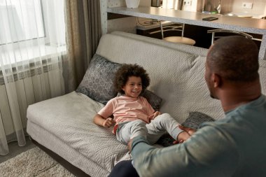 Baba, evdeki kanepede oğlunun bacaklarını tutuyor. Siyahi adam ve gülümseyen çocuk oyun oynuyor, dinleniyor, şakalaşıyor ve birlikte eğleniyorlar. Babalık ve ebeveynlik. Ev içi eğlence ve eğlence.