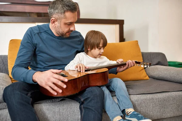 父は幼い息子に家庭でソファでアコースティックギターを教えていた 若い白人の家族関係と一緒に時間を過ごす 父親と子育て 国内レジャー — ストック写真
