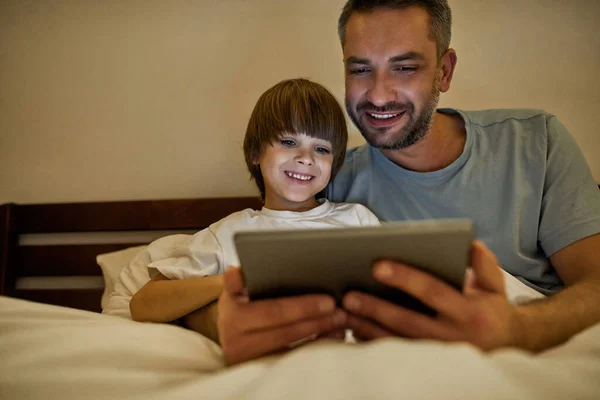 笑顔の父と小さな息子が自宅で夜にベッドでデジタルタブレットを見ています 若い白人の家族関係 父親と子育て 国内レジャー — ストック写真