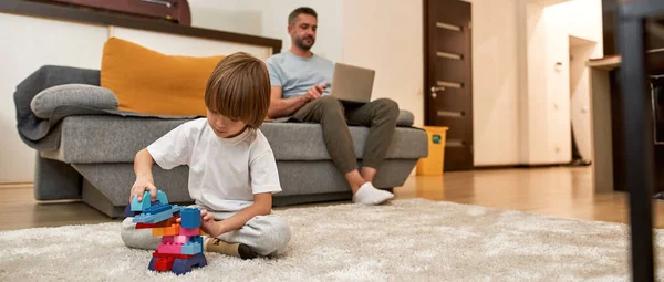 父親が自宅のソファでラップトップを使用している間 小さな男の子はカーペットの上の建設ブロックで遊んでいます 若い現代の白人の家族生活と関係 国内レジャー — ストック写真