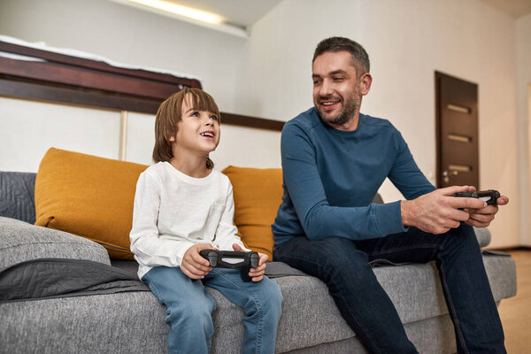 Caucasian little boy and father play video game with joysticks on sofa at home. Smiling man look at concentrated boy. Fatherhood and parenting. Family relationship. Domestic leisure and entertainment