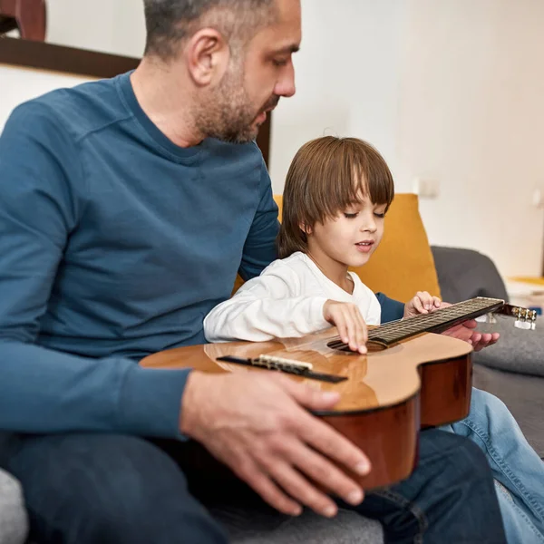 父は幼い息子に家庭のソファでアコースティックギターのコードでノートを演奏することを教えています 若い白人の家族関係 父親と子育て 国内レジャー — ストック写真