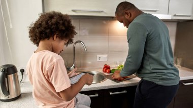 Siyah küçük çocuk dijital tablete insanları çiziyor babasının yanında ev mutfağında salata pişiriyor. Aile hayatı ve ilişki. Babalık ve ebeveynlik. Ev içi eğlence ve eğlence.