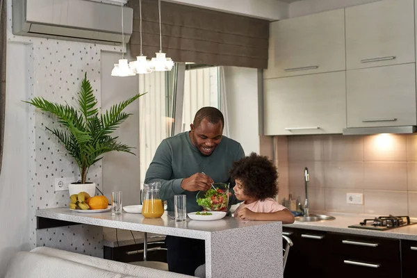 Smiling black man putting salad from bowl in plate of little son during having lunch or dinner at home kitchen. Family caring and relationship. Fatherhood and parenting. Healthy eating and lifestyle