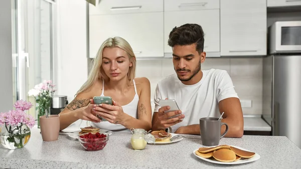 自宅のキッチンで朝食をとる際にスマートフォンを使った多人種カップルに焦点を当てた 中東の男とブロンドの白人の女の子 家庭生活 現代のアパートだ 朝の時間 — ストック写真
