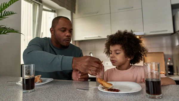 Adult father giving pizza slice to his little son during having lunch or dinner at table at home kitchen. Unhealthy eating. Young black family lifestyle and relationship. Fatherhood and parenting