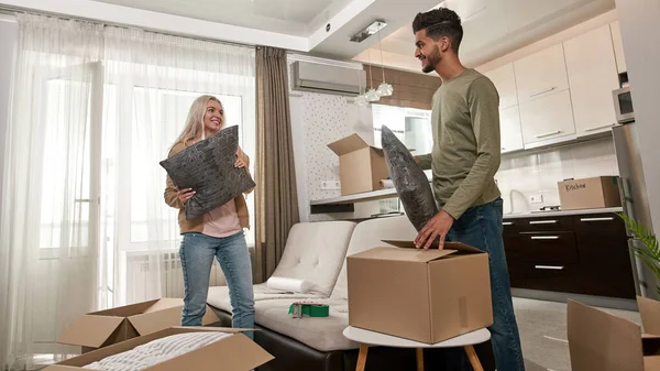 Caucasian girl and middle eastern man putting pillows in cardboard box. Young multiracial couple looking at each other and packing things for moving to new apartment. Home relocation