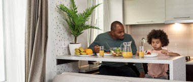 Siyah baba, öğle yemeği sırasında salata yerken ya da mutfakta yemek yerken küçük oğluna bakıyor. Aile şefkati ve ilişki. Babalık ve ebeveynlik. Sağlıklı beslenme ve yaşam tarzı