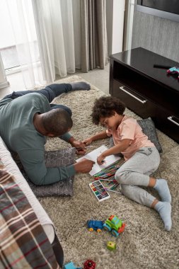 Baba ve oğulun çizim yaptığı yüksek açılı manzara. Evdeki halıdaki çizim defterindeki keçeli kalemlerle. Genç siyahi aile birlikte vakit geçiriyor. Babalık ve ebeveynlik. Ev eğlencesi, hobi ve eğlence