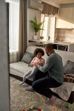 Baba evdeki kanepede oğlunu gıdıklıyor. Neşeli siyah adam ve gülümseyen çocuk oyun oynuyor, dinleniyor, şakalaşıyor ve birlikte eğleniyorlar. Babalık ve ebeveynlik. Ev içi eğlence ve eğlence.