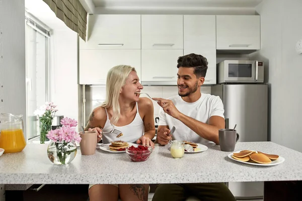 Middle eastern man feeding his caucasian girlfriend pancake during having breakfast at home. Young pleased multiethnic couple spending time together. Relationship and caring. Domestic lifestyle