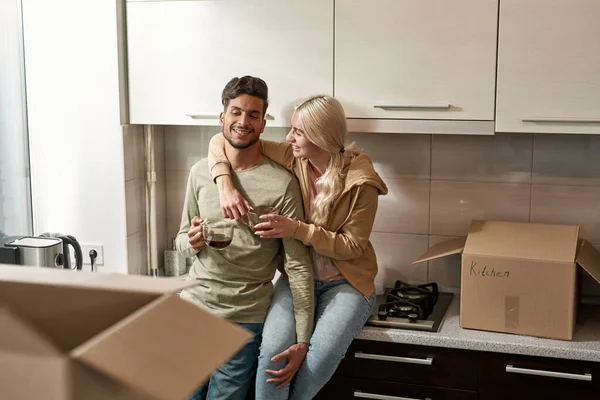 Young happy multiracial couple resting and drinking tea or coffee during packing things at kitchen. Thoughtful middle eastern man and european girl move to new apartment. Relationship. Home relocation