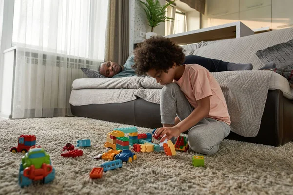 Little boy play alone with construction building blocks on carpet while his dad sleep on sofa at home. Young modern black family lifestyle and relationship. Domestic leisure, hobby and entertainment