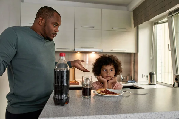 Worried dad shrugging near offended son having lunch or dinner with cola and pizza at table at home kitchen. Unhealthy eating. Young black family lifestyle and relationship. Fatherhood and parenting