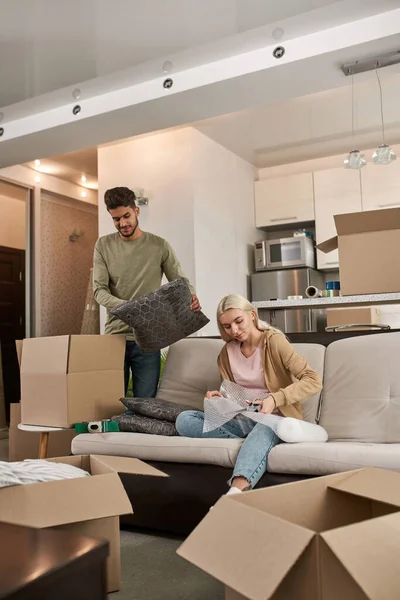 Caucasian girl cut air bubble film with scissors while middle eastern man take pillow for put in cardboard box. Young multiracial couple packing things for moving to new apartment. Home relocation