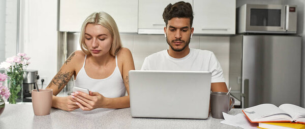 Ближневосточный мужчина работает на ноутбуке, в то время как белая женщина просматривает смартфон за столом на домашней кухне. Молодая многонациональная пара. Удаленная работа и фрилансер. Семейный образ жизни. Современная квартира