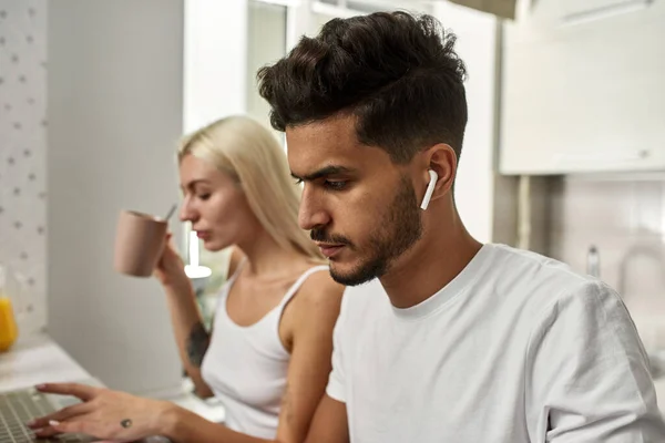 Selective focus of middle eastern man wearing earphones near blurred caucasian girl using laptop at table at home. Young multiracial couple. Remote work and freelance. Domestic lifestyle