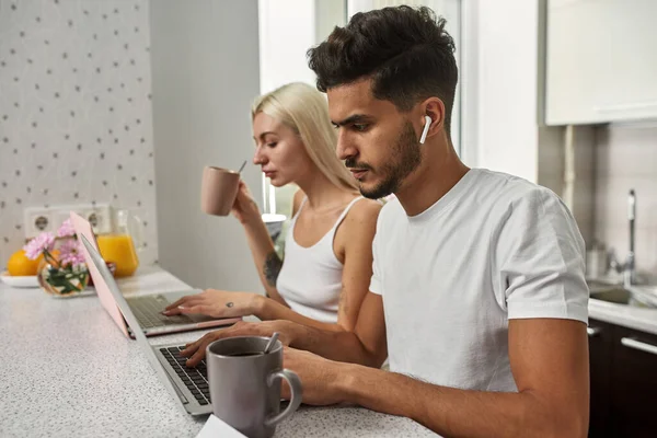 Young multiethnic couple typing and working on laptops at table at home kitchen. Middle eastern man and blonde caucasian girl. Remote work and freelance. Domestic lifestyle. Modern spacious flat